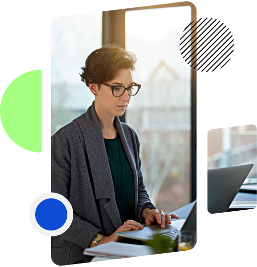 A woman wearing glasses is standing at a desk, focused on working on a laptop in a bright office setting.